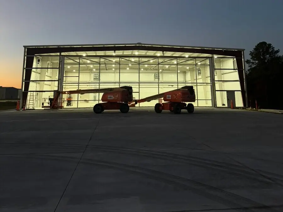 A metal building at dusk with the full-width door open and the interior lit, lifts parked on the apron