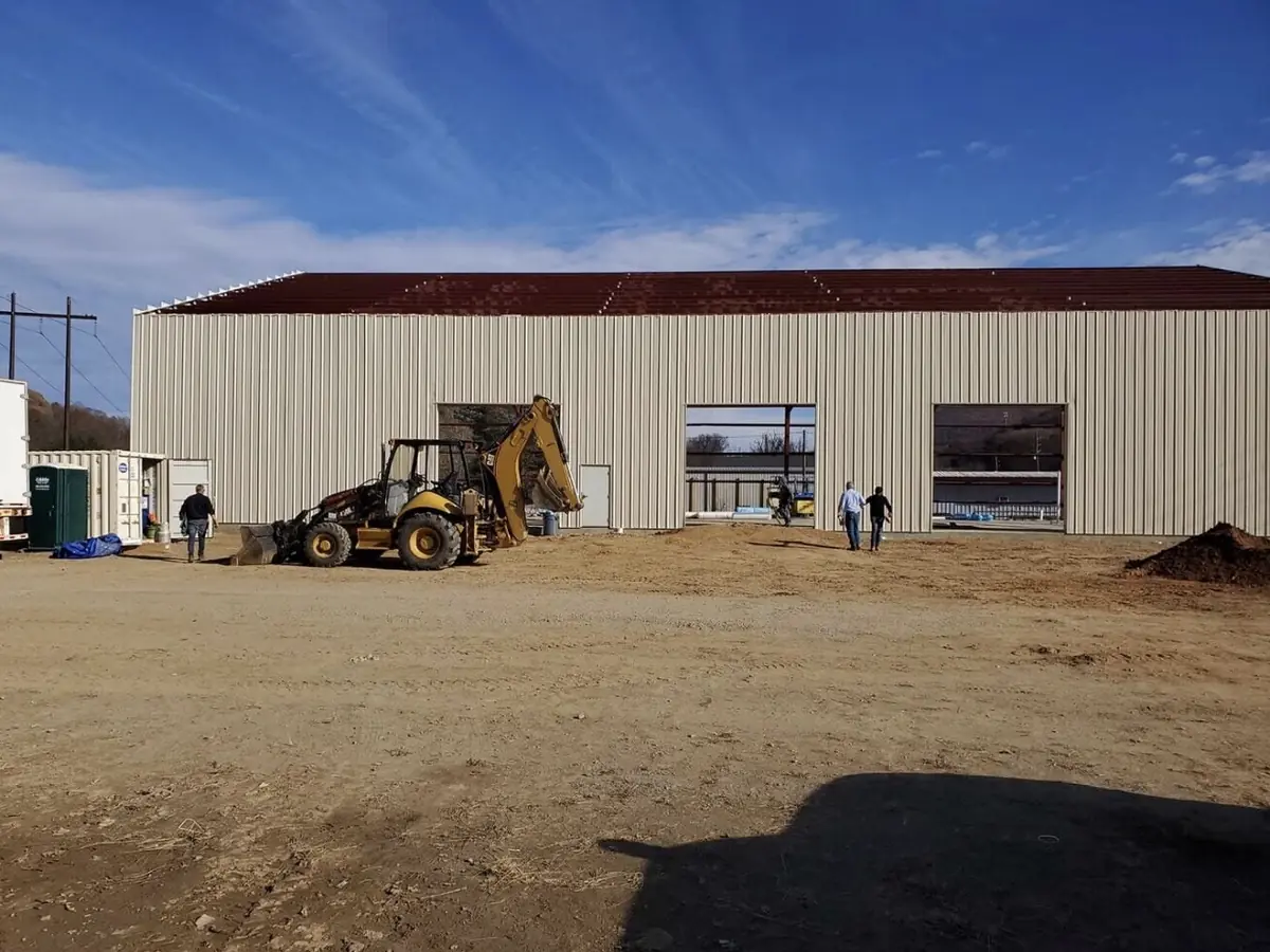 A tan metal building nearing completion with a backhoe grading the pad and crew on site
