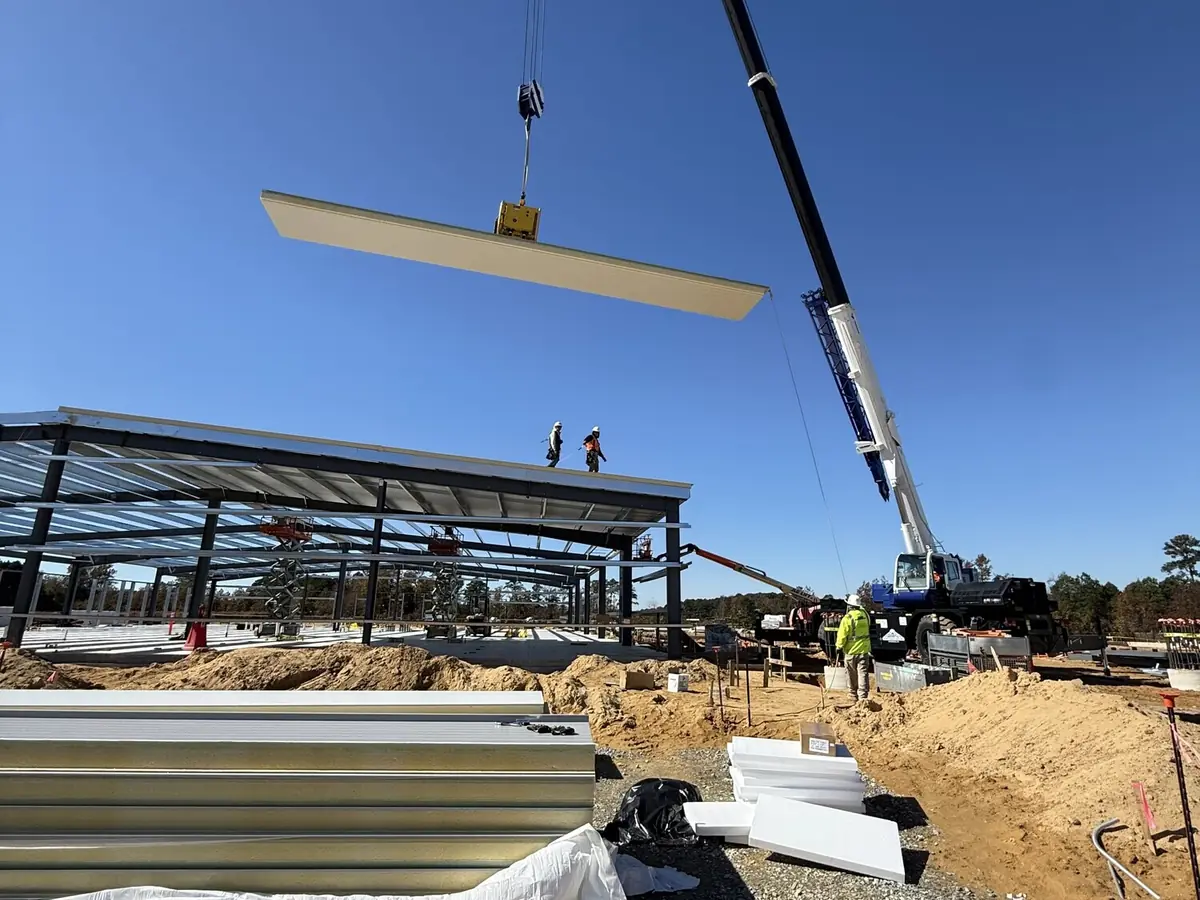 A crane lifting a steel beam into place over a partly framed metal building, crew working below
